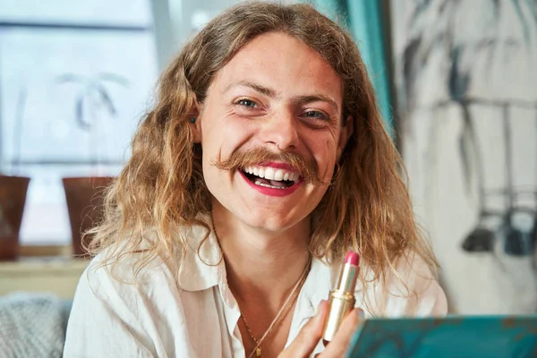 Portrait of the laughing longhaired guy with moustaches holding pink lipstick and looking at camera at home. People lifestyle concept