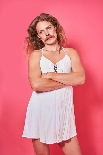 Curly longhaired man with pink lipstick at his lips and wearing creative look posing crossed arms at studio