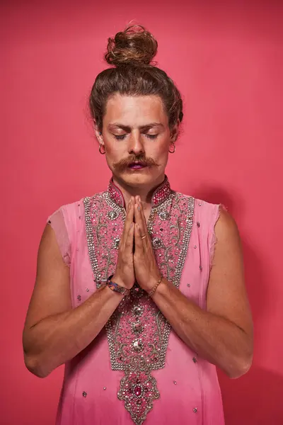 Curly longhaired man with pink lipstick at his lips wearing traditional look posing with namaste gesture at studio