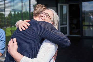 Gülümseyen son sınıf öğrencisi, evin yanında dikilirken yetişkin oğlunu kucaklıyor. İnsan ilişkileri konsepti. Stok fotoğrafı