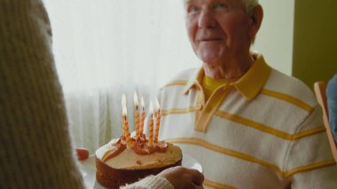 Yaşlı adam doğum günü pastasına bakıyor ve mum üflüyor. Yaşlı çift evde doğum günü partisi veriyor.