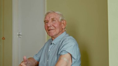 İhtiyarlar için aşı. Doktorun ofisinde oturan ve koluna yapışkan alçıyı 19 aşıdan sonra gösteren mutlu kıdemli adam.