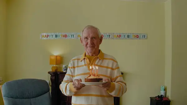 Old man holding birthday cake and blowing candles. Elderly couple having birthday party at home