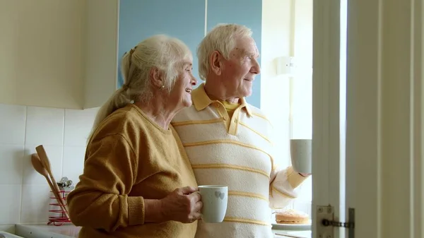 Happy senior couple drinking coffee or tea on home kitchen. Happy elderly spouses looking at window together