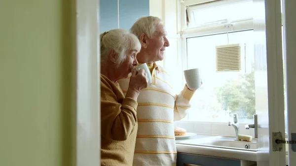 Happy senior couple drinking coffee or tea on home kitchen. Happy elderly spouses looking at window together