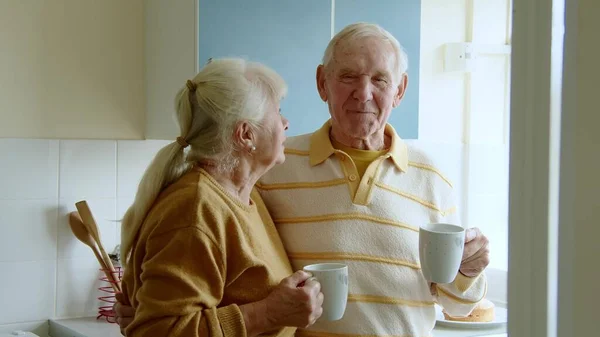 Happy attractive senior couple in home wear standing at white cozy kitchen, drinking fresh morning coffee together, enjoying good morning, sharing plans for day