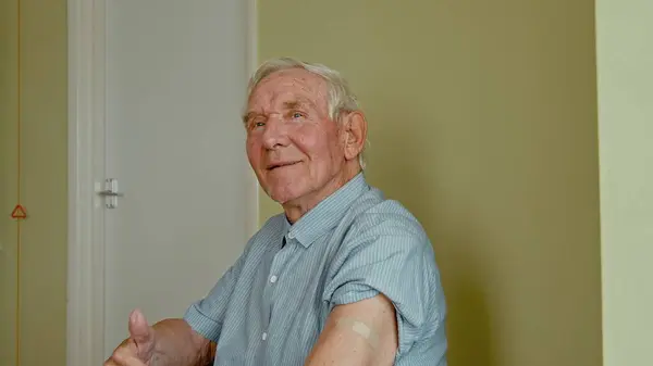 Vaccination for elders. Happy senior man sitting in the doctors office and showing his arm with adhesive plaster after covid 19 vaccine