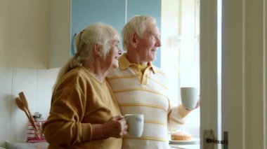 Ev mutfağında çay ya da kahve içen mutlu bir çift. Mutlu yaşlı eşler birlikte pencereye bakıyor.