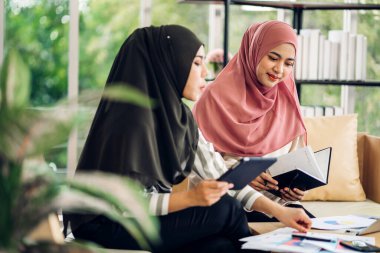 Gülümseyen mutlu Asyalı güzel Müslüman kadın dizüstü bilgisayarla rahatlıyor, çalışıyor ve analiz ediyor. Başlangıç projesi planı ve evdeki beyin fırtınası ile strateji tartışıyorlar.