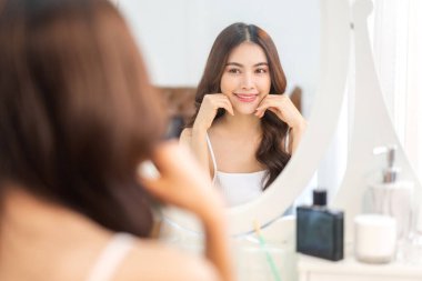 Genç, güzel Asyalı bir kadının, temiz, sağlıklı, beyaz bir tenle aynaya bakması. Asyalı bir kızın elleriyle yüzüne dokunması ve evde krem sürmesi. Spa ve güzellik konsepti.