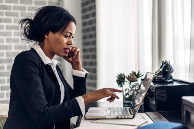 Gülümseyen mutlu Afro-Amerikan siyahi kadının portresi masada otururken dizüstü bilgisayar teknolojisi kullanarak rahatlıyor. Ev konseptinde çalışan yaratıcı Afrikalı genç bir kız.
