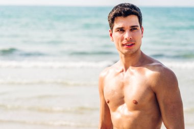 Gülümseyen yakışıklı seksi adamın portresi kaslı fit vücudunu gösteriyor tropik sahilde durup eğleniyor ve rahatlıyor.