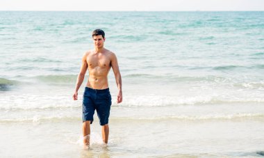 Gülümseyen yakışıklı seksi adamın portresi kaslı fit vücudunu gösteriyor tropik sahilde durup eğleniyor ve rahatlıyor.