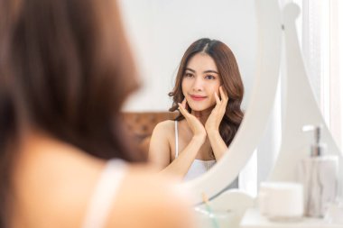 Genç, güzel Asyalı bir kadının, temiz, sağlıklı, beyaz bir tenle aynaya bakması. Asyalı bir kızın elleriyle yüzüne dokunması ve evde krem sürmesi. Spa ve güzellik konsepti.