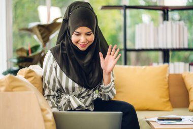 Genç gülümseyen mutlu Asyalı Müslüman kadın dizüstü bilgisayarla rahatlıyor. Çalışıyor ve analiz ediyor. Başlangıç projesi planı ve evdeki beyin fırtınasıyla strateji tartışıyorlar.