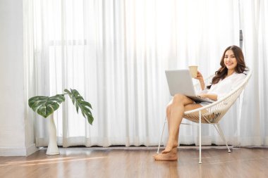 Genç gülümseyen mutlu Asyalı kadın evdeki yatak odasında dizüstü bilgisayar kullanarak rahatlıyor. Ev konseptinden çalışıp klavyede yazan genç yaratıcı kız.