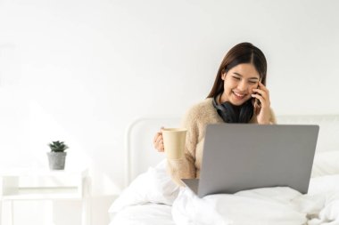 Genç gülümseyen mutlu Asyalı kadın evdeki kanepede dizüstü bilgisayarla rahatlamak için çalışıyor. Yaratıcı genç kız çalışıyor ve ev konseptinden akıllı telefon kullanıyor.