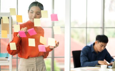Gündelik yaratıcı mutlu iki gülümseme Asyalı iş adamları strateji analizi ve beyin fırtınası planlar. Modern ofisteki cam panoda yeni bir proje başlatma fikrini paylaşmak için çıkartma notu yapıştırırlar.
