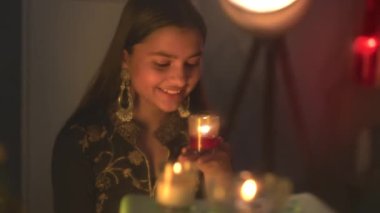 Gülümseyen, büyük küpeler takan, elinde kırmızı camla oturan, Diwali 'de mum yakan güzel bir kız. Dekoratif çevreyle Diwali festivalinin tadını çıkaran kız