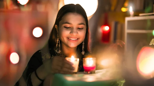 Smiling pretty Hindu girl wearing big earrings sitting look hold burning glass candles closer to face, Indian teen enjoy Diwali or Deepavali festival by lightening Diyas with colorful background