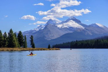 A canoe heads out onto glacier-fed Maligne Lake in Jasper National Park with majestic mountains and a glacier in the background and a bright blue sky with white clouds. 