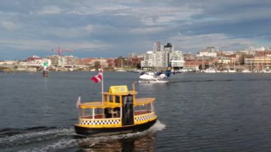 A twin-engine propeller seaplane and yellow water taxi cross paths in Victoria's busy inner harbour with the city skyline and Johnson St. bridge in the background. 