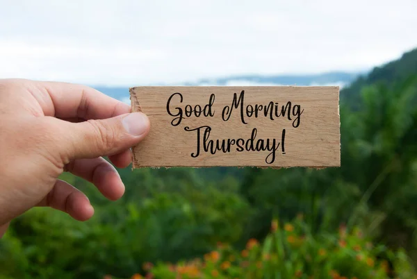 Hand holding wooden banner with Good Morning Thursday text. With beautiful nature background.