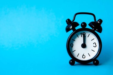 Black alarm clock isolated on blue background. The clock set at 12pm. Copy space. clipart