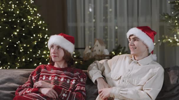 新年の雰囲気 10代の子供たちがソファに座ってテレビを見ています クリスマスツリーライトが背景に輝きます クリスマス休暇の概念 — ストック動画