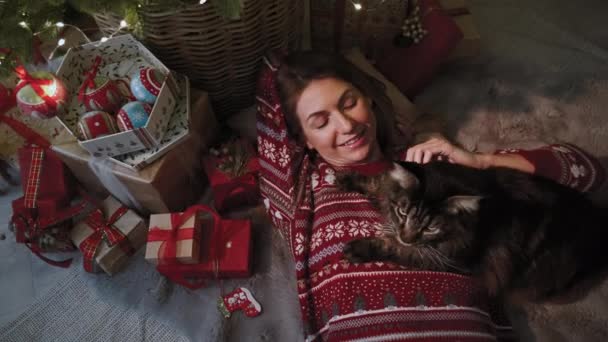 新年の服を着た美しい女の子は 彼女の好きな猫の会社のクリスマスツリーの下にあります カメラを離して 新年のお祝いの概念 — ストック動画