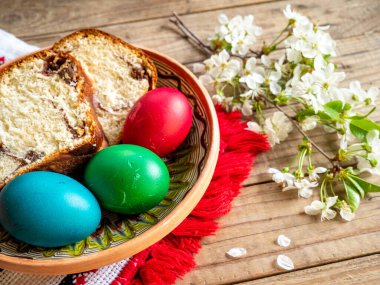 Hasır bir sepette renkli Paskalya yumurtaları ve ahşap arka planda elma çiçekleriyle birlikte tatlı ekmek dilimleri.