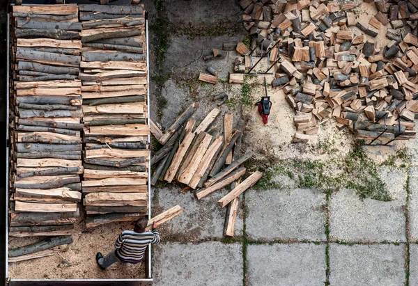 An unrecognized person unloads firewood from a truck. Chainsaw set down by cut logs in a backyard. A bird\'s-eye view.
