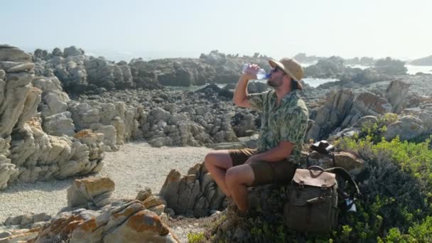 夏休みは海辺でリラックスした時間 放浪者南アフリカの喜望峰で石の上にポーズをとる旅行者の男 海の海岸で息をのむような景観自然景観を楽しむ藁帽子の男 — ストック動画