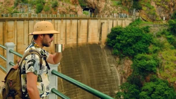 山の峡谷にある水力発電所です 山の川の水力発電所が湖に水を放出します 男性旅行者はマグカップから熱いお茶を飲みダムの橋の上に立ち — ストック動画