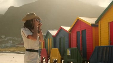 Safari şapkalı bir kadın gezgin, Cape Town, Güney Afrika 'daki renkli hamamböcekleriyle Cape Town' daki St. James Plajı 'ndaki renkli soyunma odalarının arka planında fotoğraf çekiyor..