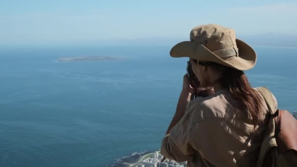 サファリの服と帽子の旅行者は高い山から海の写真を撮る ブーツとショートパンツのハイカーが峠を登り山の写真を撮りました カメラを持った写真家 — ストック動画