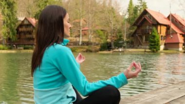 Göl kenarındaki dağlarda yoga yapan atletik bir kadın. Kadın doğada yoga yapıyor. Lotus pozisyonunda çimenlerde oturan ve şehir parkında büyük ağaçlarla el kaldıran kadın.