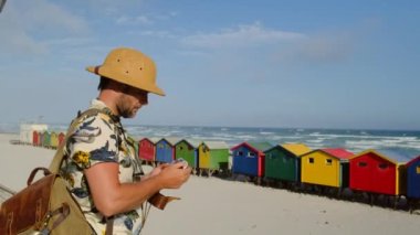 Erkek gezgin Afrika 'da gezer, fotoğraflar. Renkli soyunma odalarının yanındaki okyanus manzarasını seviyor. Kumsalda renkli banyo kulübeleri. Cape Town 'da Muizenberg sahilinde siyahi evler - Güney Afrika 