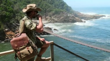 Şapkalı ve sırt çantalı bir erkek yolcu, okyanustaki asma köprünün yanındaki bir bardaktan sıcak çay içiyor. Ateş, çadır ve yakacak odunları olan bir turizm kampı. Seyahat, macera.,