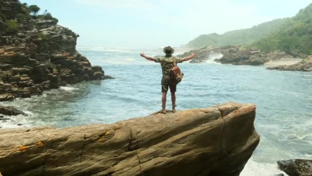 男の旅人が海と波の上の石の上に立って吊橋の近くの停止点で両手を横に上げている テントや薪の観光キャンプ — ストック動画