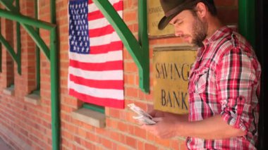 Banka veznedarı dolar banknotlarını sayıyor. Banka veznedarı ellerinde dolar banknotları, mali ve yatırım kavramlarını sayıyor. Şapkalı bir adam banka kapısının yanında oturur ve Amerikan dolarlarını sayar.