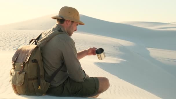 ネバダ砂漠の山の上で黄金の夕日を見ている男 サファリの帽子とシャツを着た男性が砂漠の砂の上に座って助けを求めています 砂漠で喉の渇きと飲料水を感じる — ストック動画