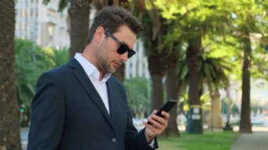 Genç sıradan bir iş adamı cep telefonuyla mesaj yolluyor. İş, yaşam tarzı, teknoloji ve sosyal medya ağı kavramı. Takım elbiseli bir adam elinde telefonla sokakta dikiliyor.