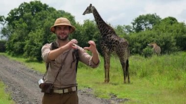 Keşif giysileri içinde gezen bir adam kalbi Savannah 'da zürafaya gösteriyor. Güneş şapkalı bir erkek ve savanadaki yetişkin bir zürafanın arka planına karşı seyahat ediyor. Safariye çıkan insanlar