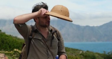 Yorgun bir gezgin alnından terleyerek siliyor. Erkek gezgin okyanusun yakınındaki bir dağ uçurumunda duruyor. Serbest çalışan genç bir gezgin bir yerlerde şapka takıyor ve dağ manzarasının tadını çıkarıyor.