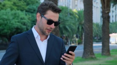 Takım elbiseli şık ve başarılı bir adam elinde telefonla sokakta duruyor. Parkta kol saatine bakan tanınmayan bir adam. Resmi takım elbiseli bir erkek dışarıda zamanı kontrol ediyor. Takım elbiseli yakışıklı bir adam.