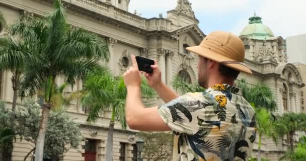 一个穿着旅行服装的旅行者在巴塞罗纳市中心的手机上拍照 美国男性旅行者在公园里坐着休息 度假时欣赏建筑 欧洲的男性旅行者A — 图库视频影像