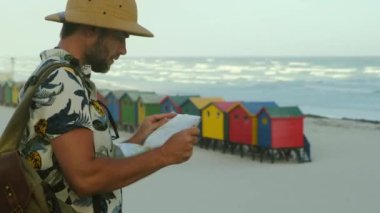 Erkek gezgin Afrika 'da seyahat eder ve okyanustaki haritaya bakar. Cape Town, Güney Afrika 'daki ünlü St. James plajının renkli soyunma odaları. Sahilde renkli ahşap kabinler