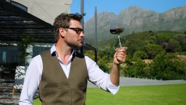 幸せな成功した男性のワインメーカーは味を試飲し ワインセラーに透明なガラスに注がれた赤ワインの品質を確認しています 白いシャツを着たイタリア人がワインを片手にテーブルに座り — ストック動画