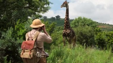 Safaride fotoğraf çeken bir adam Afrika savanasında yaprakları kuru mevsimde kızıl toprakla yiyen vahşi bir zürafa. Safari şapkalı bir erkek gezgin zürafanın fotoğrafını çekiyor.